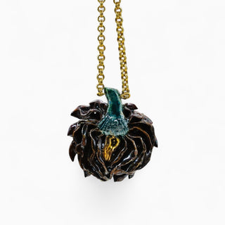 A close-up of a dark brown artichoke pendant necklace with a glossy teal stem and gold accents, suspended from a gold chain against a plain white background.