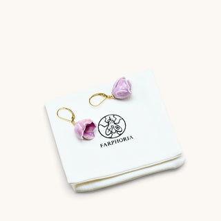 A pair of dangling earrings with light purple flower-shaped charms and gold hooks, displayed on a white jewelry pouch printed with the brand name FARPHORIA.