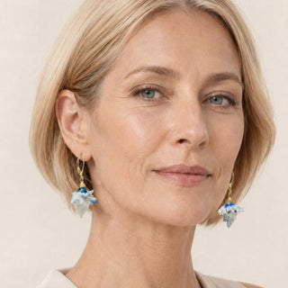 A close-up portrait of a woman with short blonde hair wearing gold dangle earrings with light blue and white porcelain earrings made from real thistle by FARPHORIA London